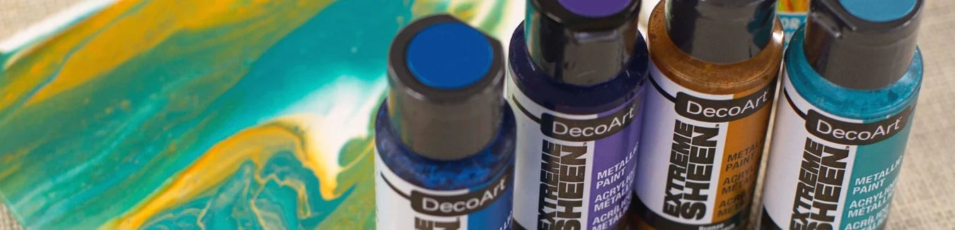 A vibrant collection header banner featuring an array of DecoArt Extreme Sheen metallic paint bottles showcasing shimmering gold, bronze, and silver hues on a rustic wooden craft table next to beautifully finished projects.