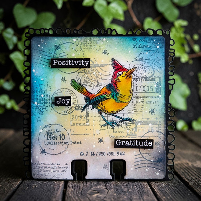 AALL & Create A7 Acrylic Stamp – Song from Torino Decorative card with a colorful bird illustration and motivational words on a wooden surface with greenery in the background.